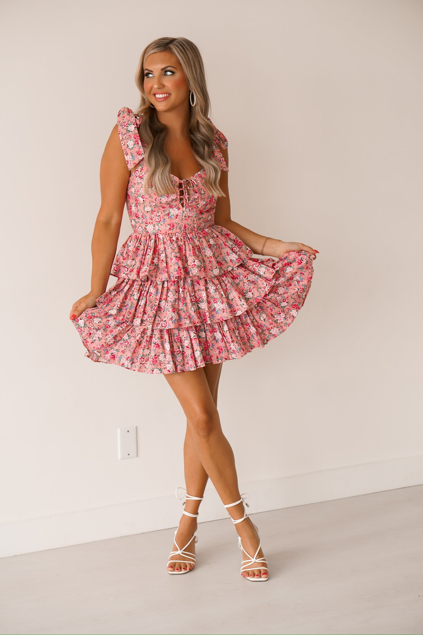 Blonde headed women standing against a white wall wearing a pink floral dress with white heels