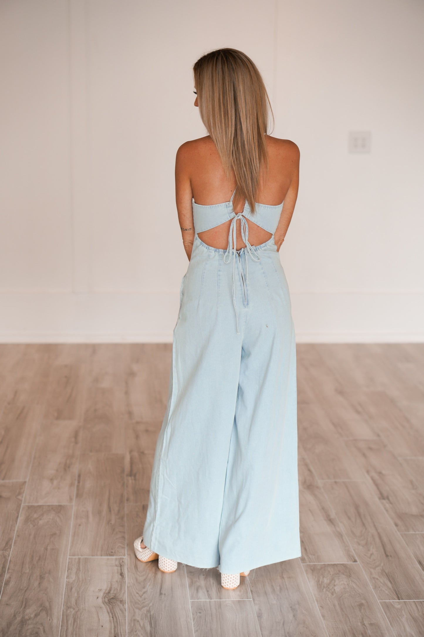 showing off the back of the long denim jumpsuit that has an open back and zipper