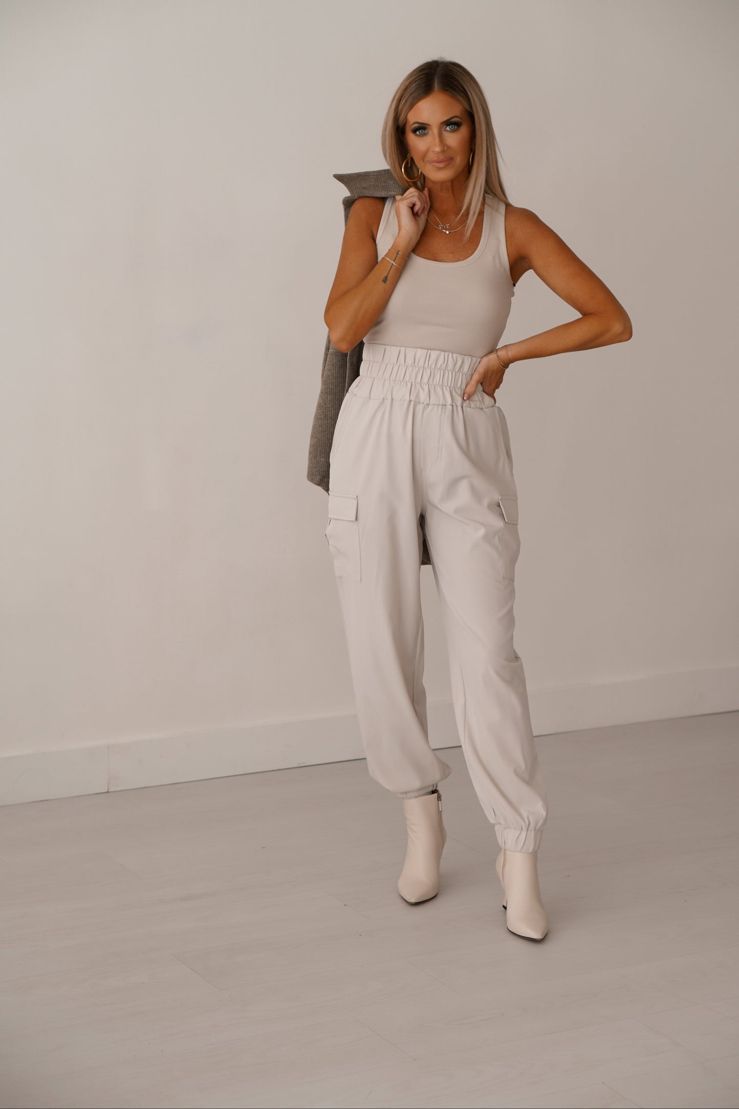 Blonde girl wearing cream cargo joggers holding green shacket over shoulder. She is standing in front of white wall.