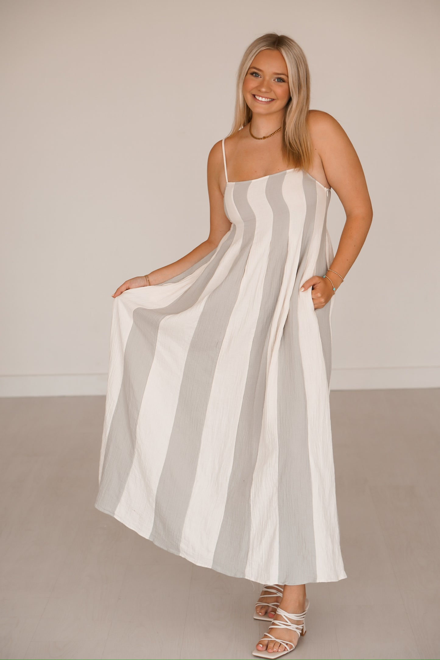Blonde headed lady standing against a white wall wearing a blue and cream striped dress that has pockets