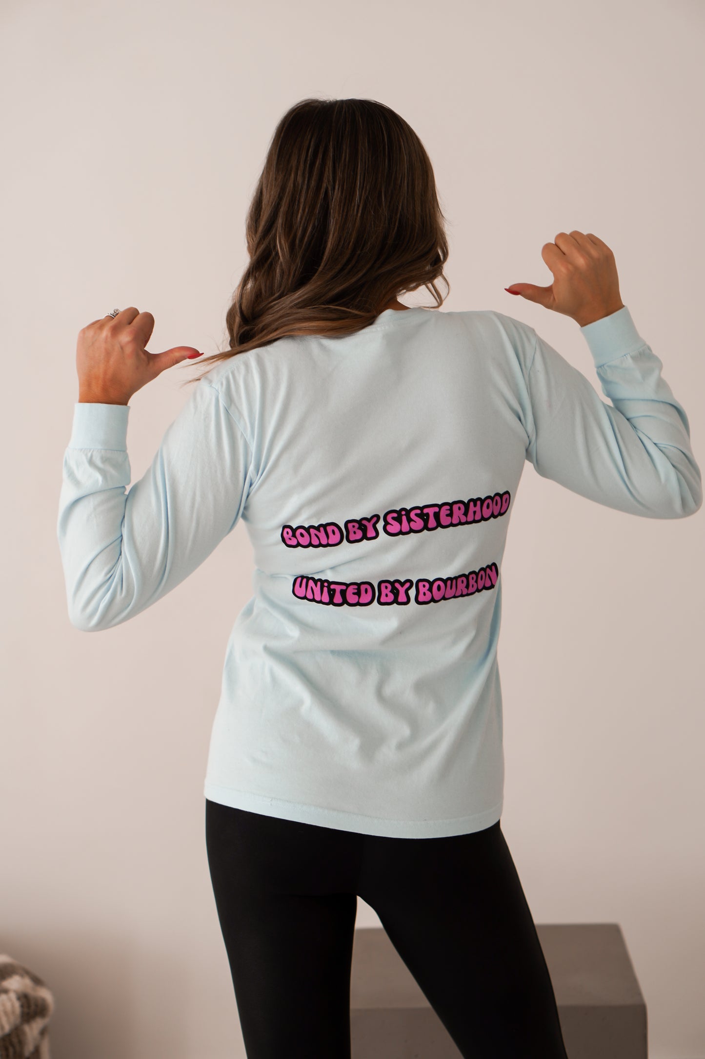 Girl standing in front of white wall showing back of blue long sleeve unisex shirt with pink writing that says bond by sisterhood united by bourbon. 