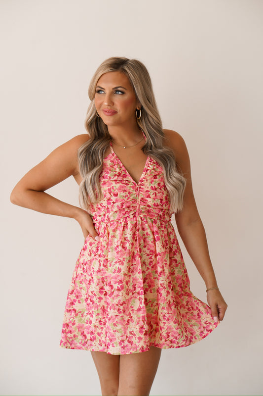 blonde headed girl standing against a white wall, wearing a short floral halterdress. 