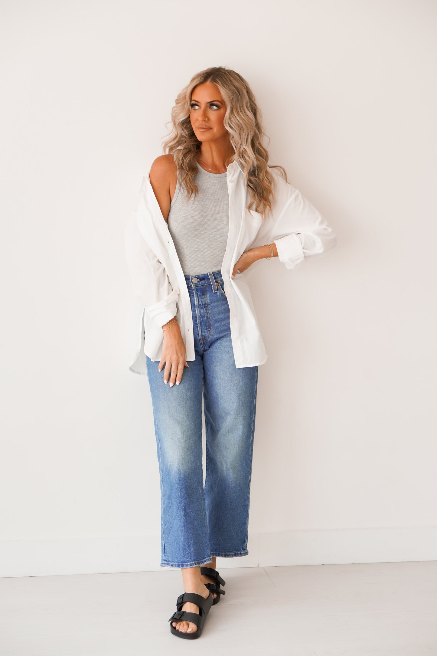 BLonde headed women standing against a white wall wearing black sandals, blue jeans, and a grey crop top with a white button up over it