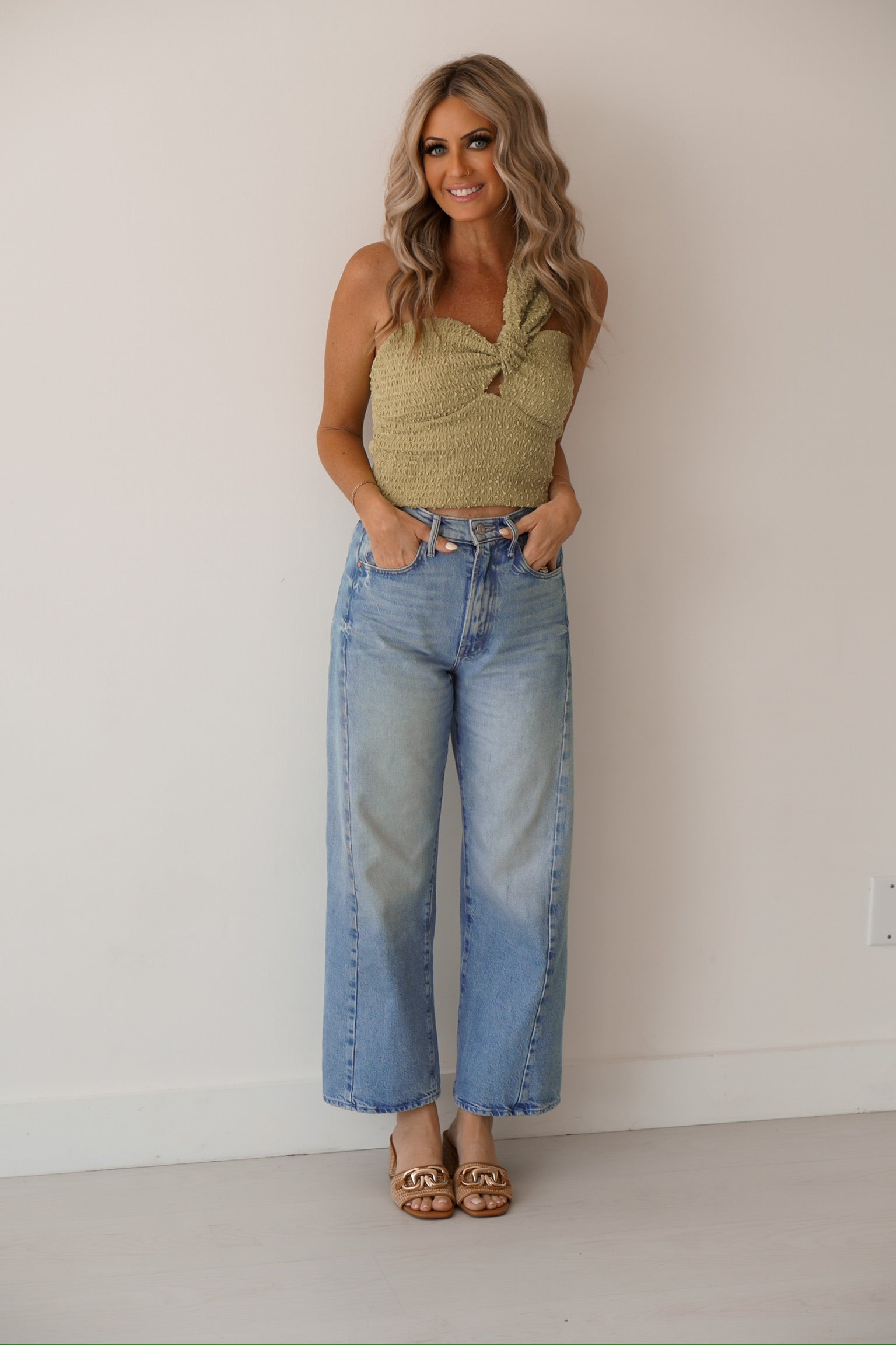 blonde headed girl standing against white wall wearing a lime green one shoulder top, high waisted jeans, and some tan sandals
