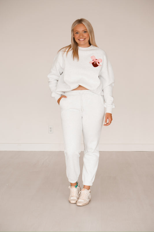 Blonde headed girl standing against a white wall wearing grey sweatpants and a grey sweatshirt that has a football and pink bow on it