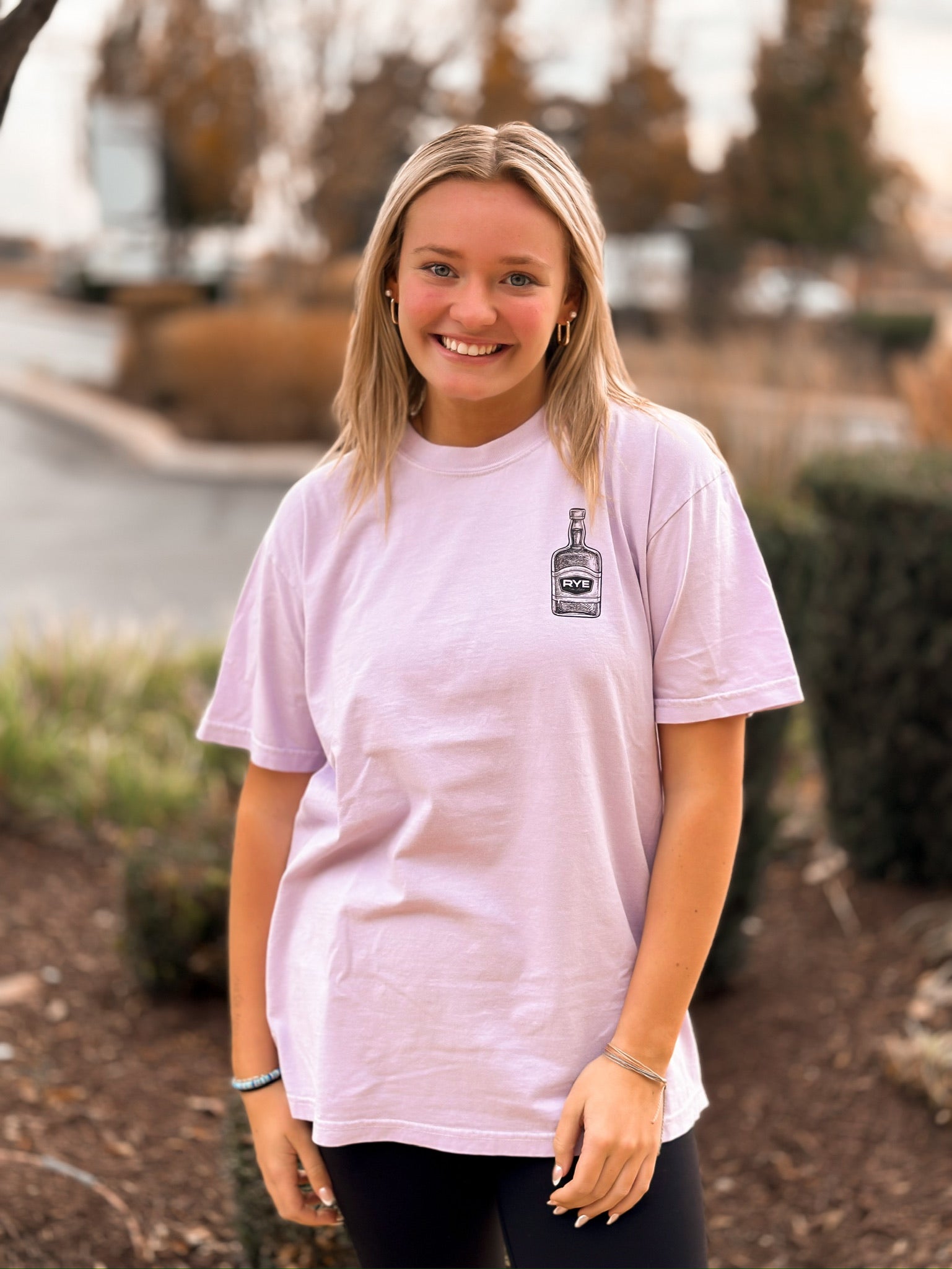 Blonde girl wearing purple short sleeve shirt with RYE bourbon bottle standing in front of trees. 