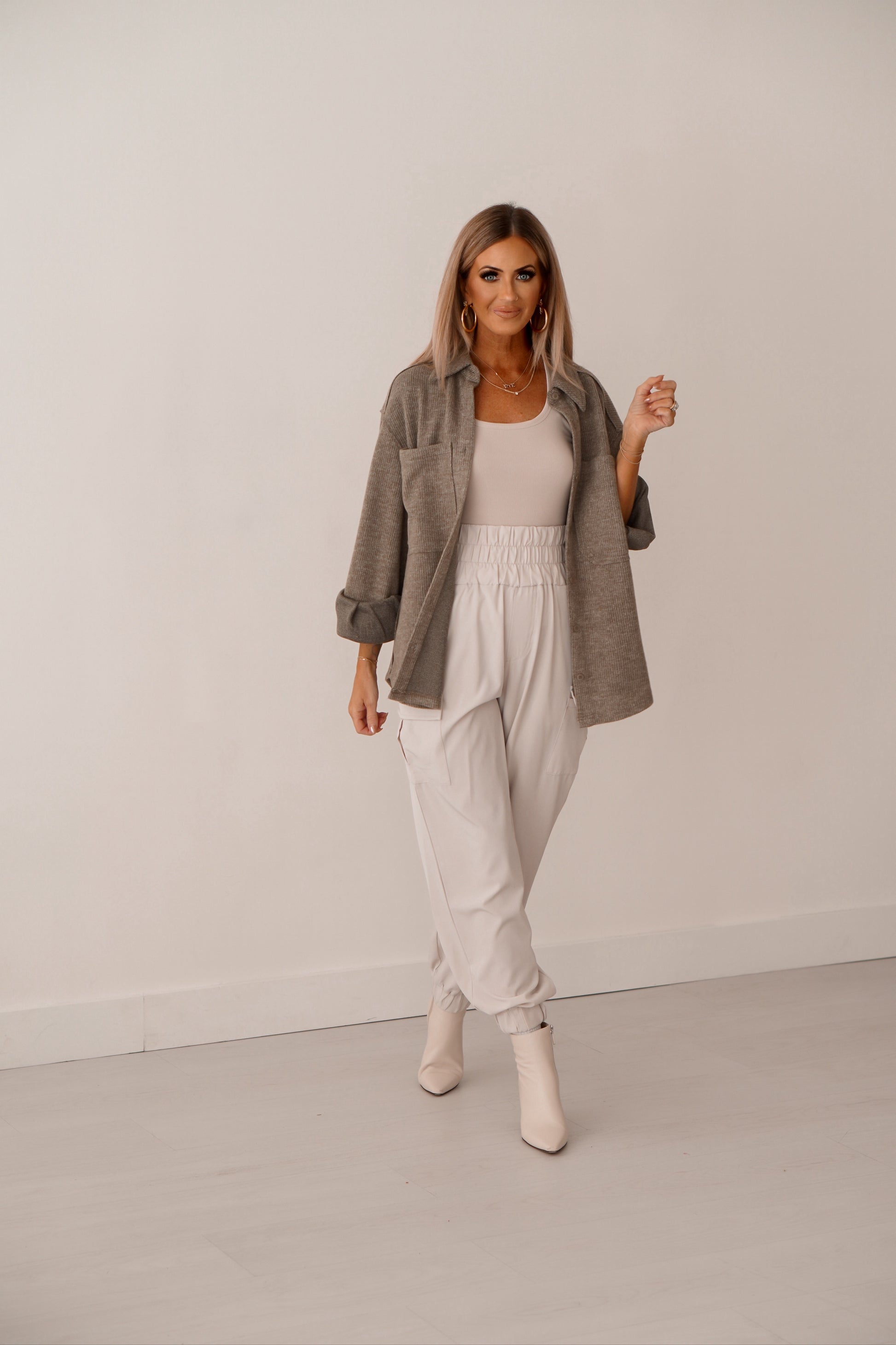 Blonde girl wearing a cream tank, cream joggers and olive green shacket. She is standing in front of white wall. 