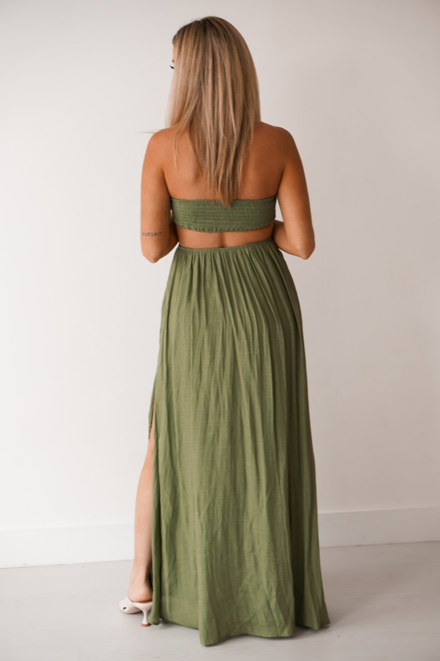 girl standing against a white wall wearing a long green dress that has cutouts on the side of the hips. SHe is wearing sandals. She is showing off the back of the dress which is open on the lower back 