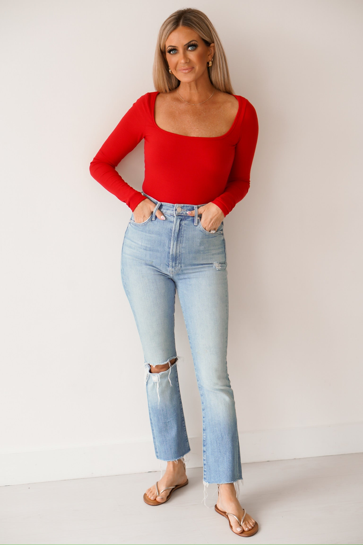Blonde headed girl standing against a white wall wearing sandals, denim jeans, and a red long sleeve top 