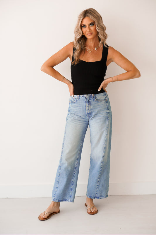 girl standing against a white wall wearing a black tank top, high rise denim jeans, and sandals