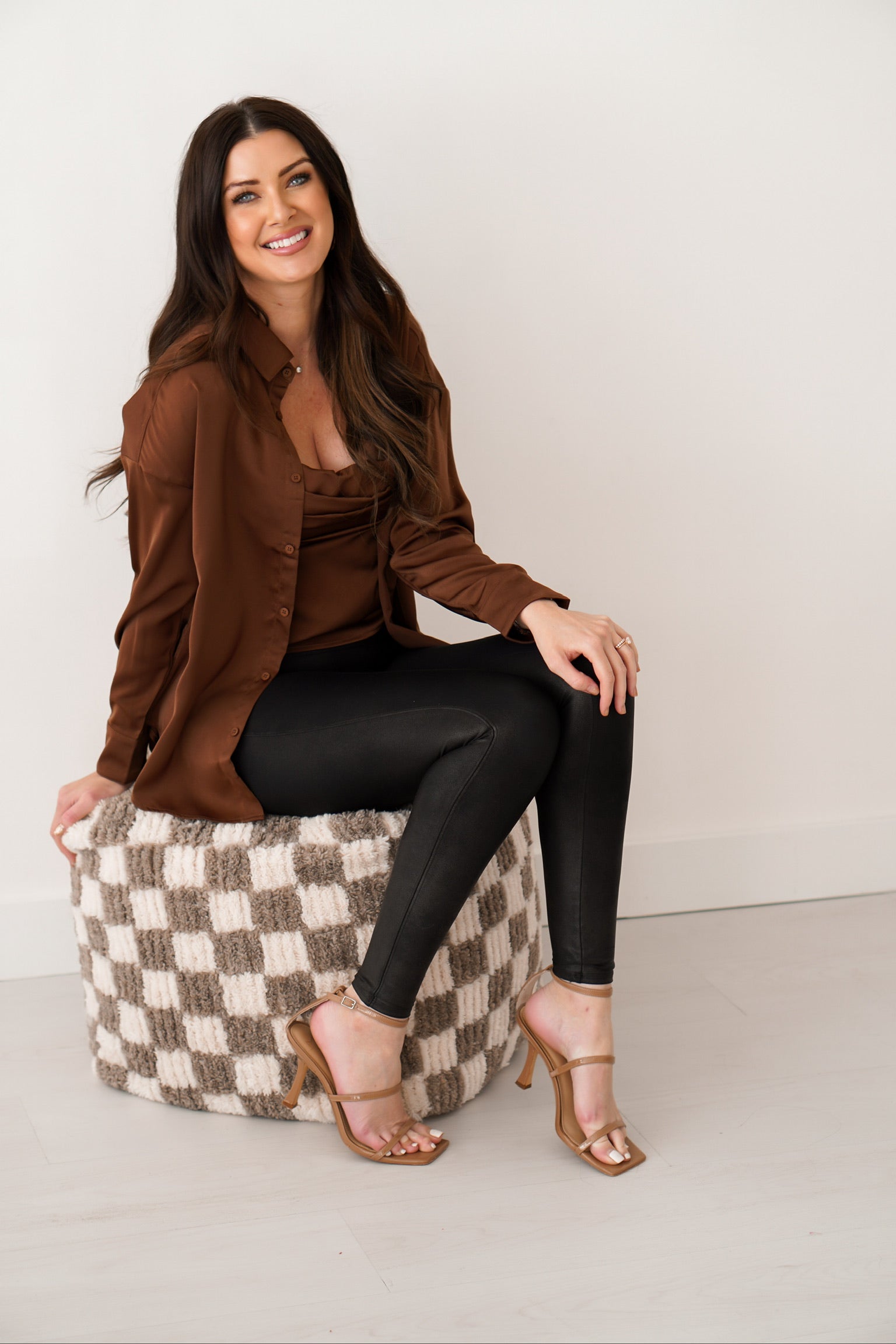 Brunette girl sitting on checkered ottoman wearing chocolate brown silk top set in front of white wall. 