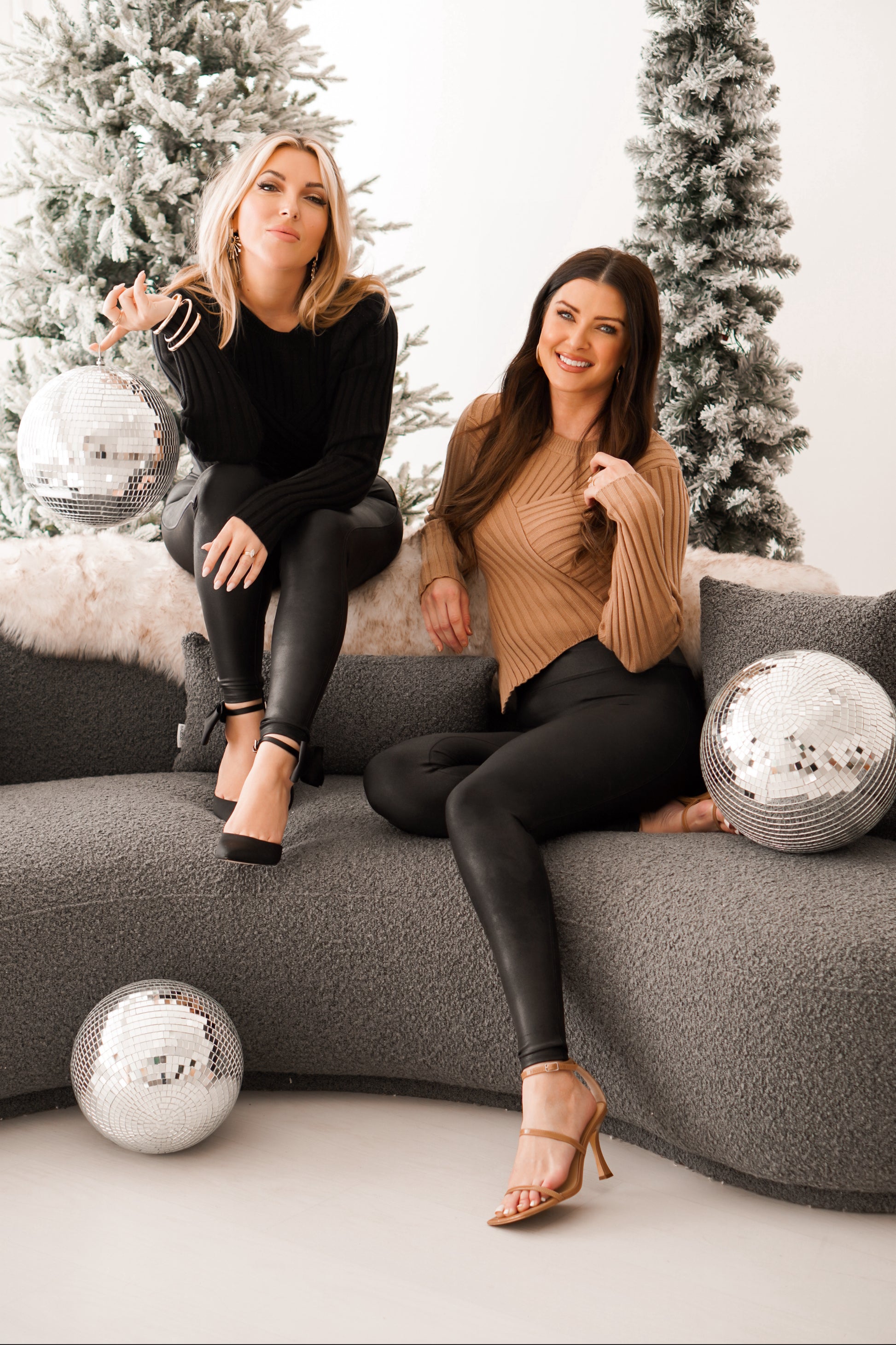 Blonde girl wearing black sweater with V detail in front with Brunette girl wearing same sweater top with V detail on the bottom in tan. They are both sitting on gray couch with disco balls. 