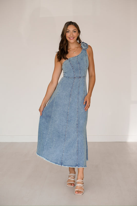 Brunette standing against a white wall wearing a long denim one shoulder dress 