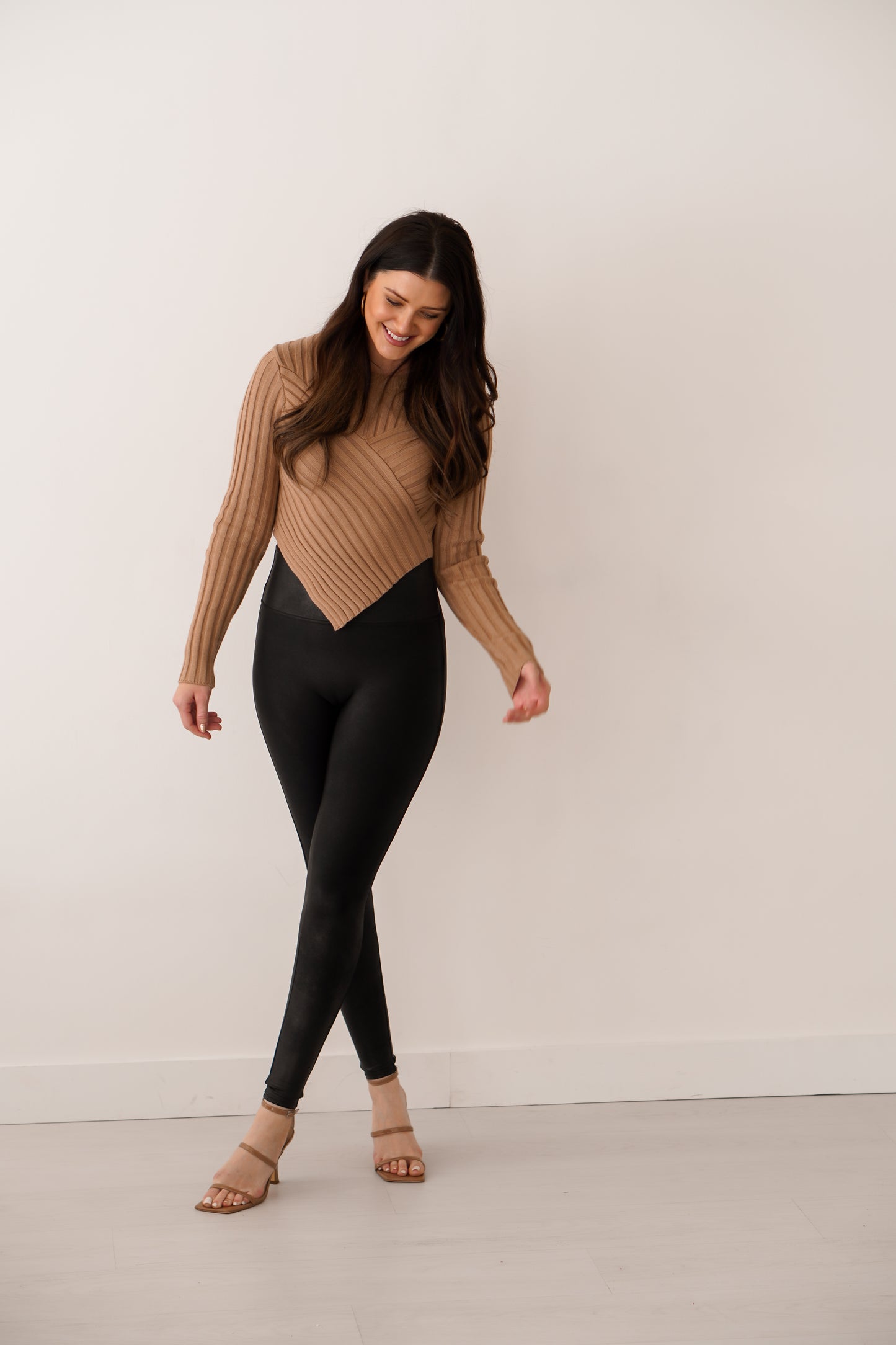 Brunette girl wearing V tan sweater with black leggings with legs crossed. 