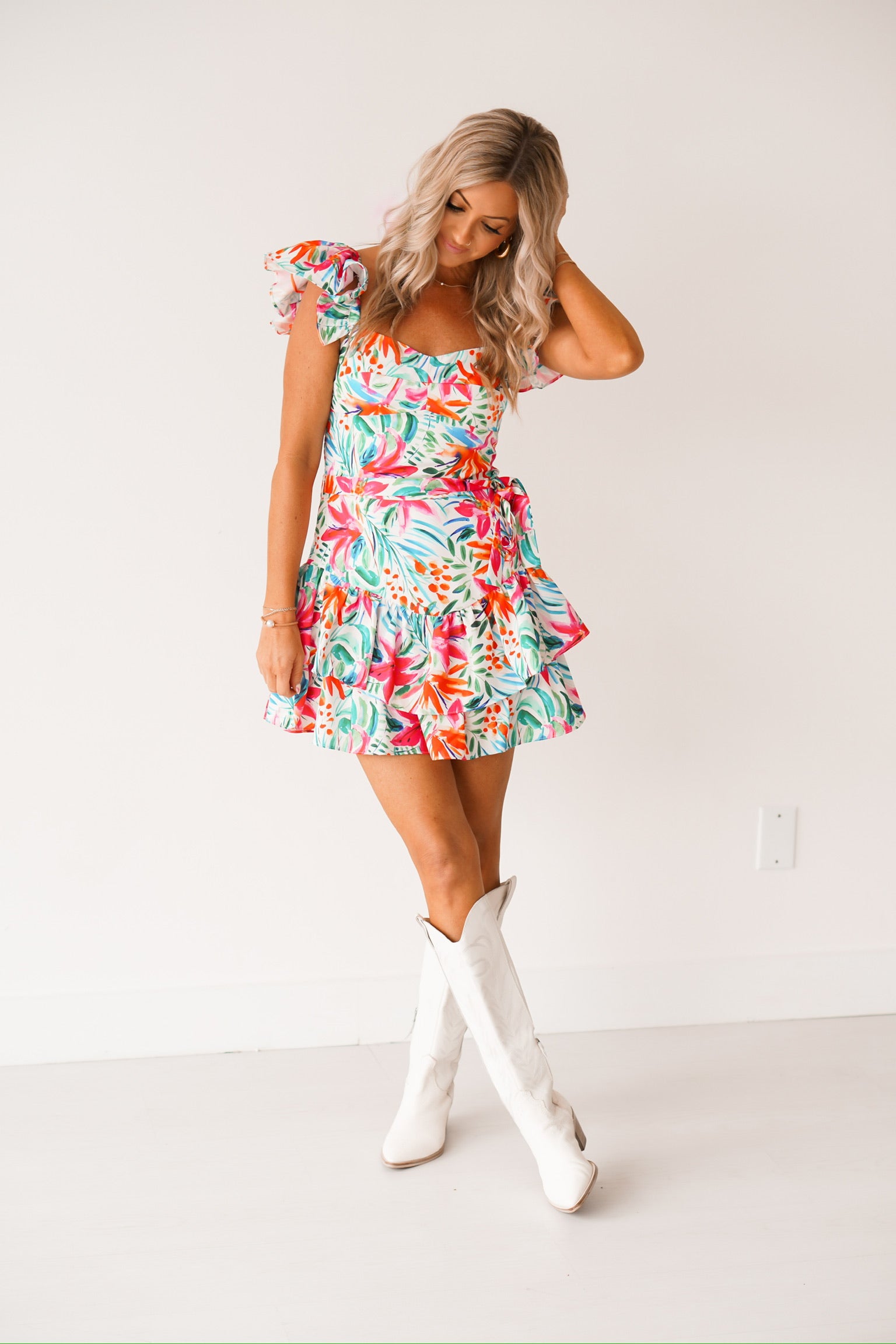 Blonde headed girl standing against a white wall wearing cowgirl boots and a tropical print romper