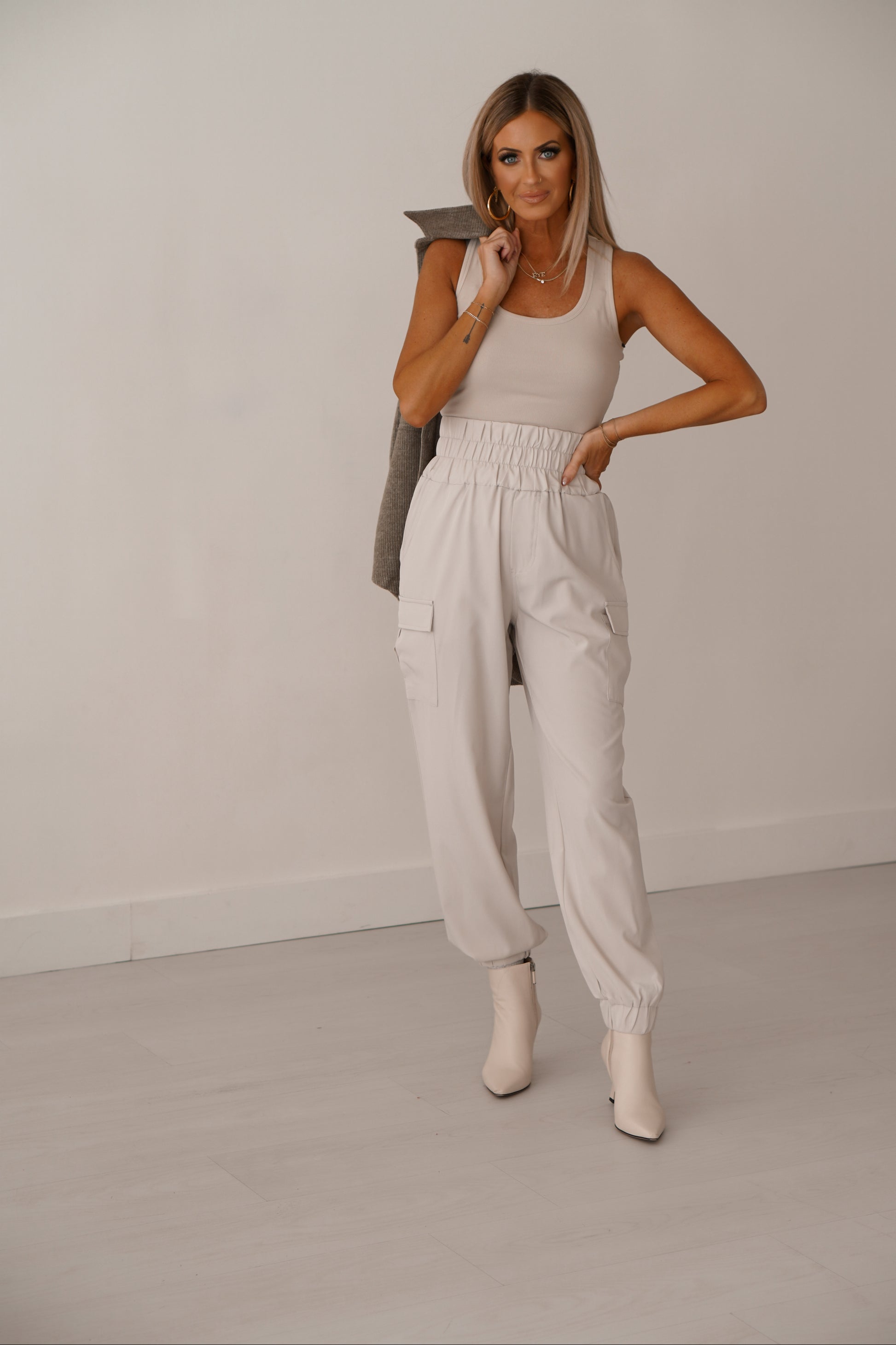 Blonde girl wearing cream cargo joggers holding green shacket over shoulder. She is standing in front of white wall. 