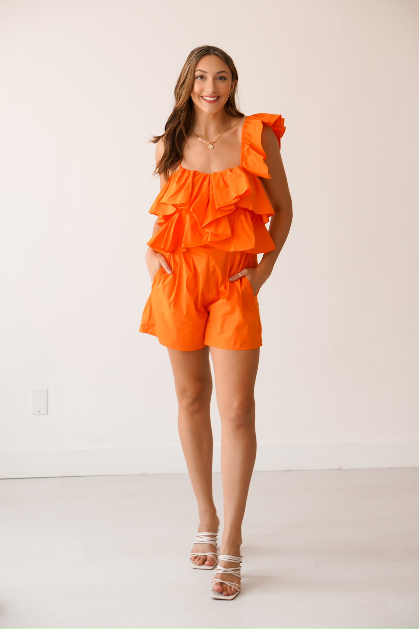 Brunette standing against a white wall wearing a bright orange one shoulder romper 