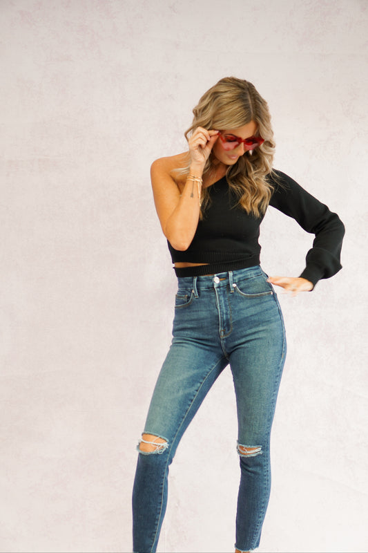 girl looking down at her shirt that is a black one shoulder sweater with cut out detail on waist wearing jeans and heart shaped glasses