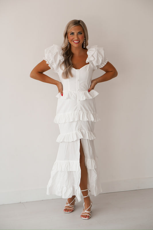 blonde headed women standing against a white wall wearing a white ruffled maxi dress that has a slit in it