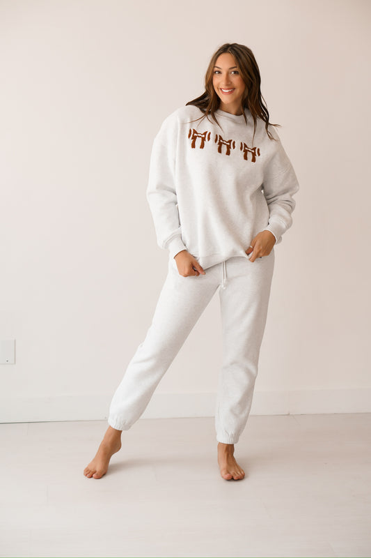 Brunette headed girl standing against a white wall wearing light grey sweatpants and a light grey sweatshirt with a football shaped in a bow on it