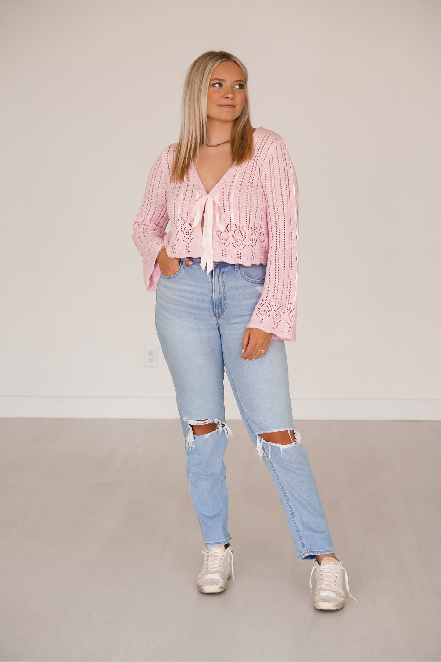 Blonde headed lady standing against a white wall wearing denim jeans and a light pink crocheted top