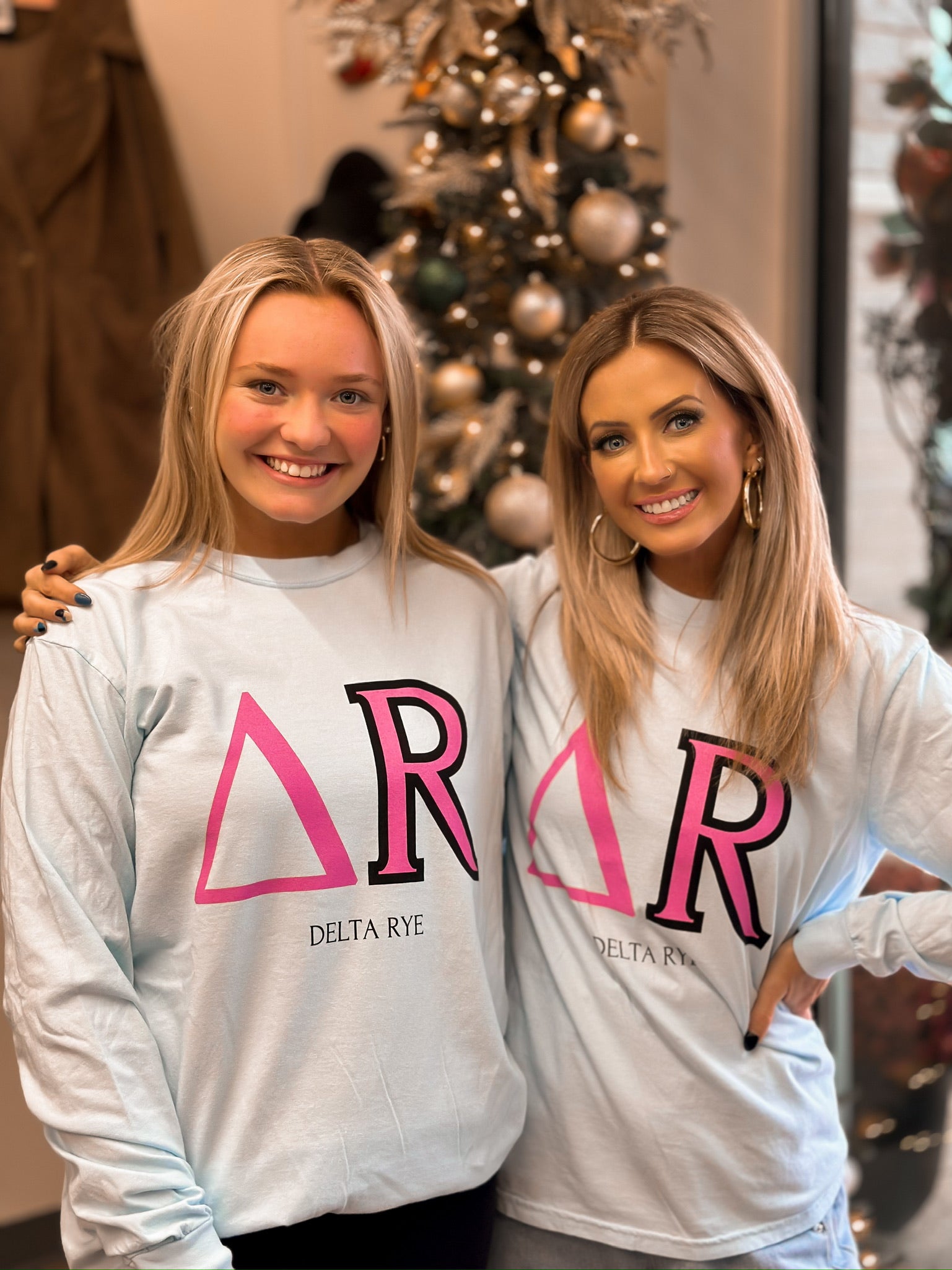 Two blond girls wearing a blue long sleeve unisex shirt with pink and black greek letters for Delta RYE. 