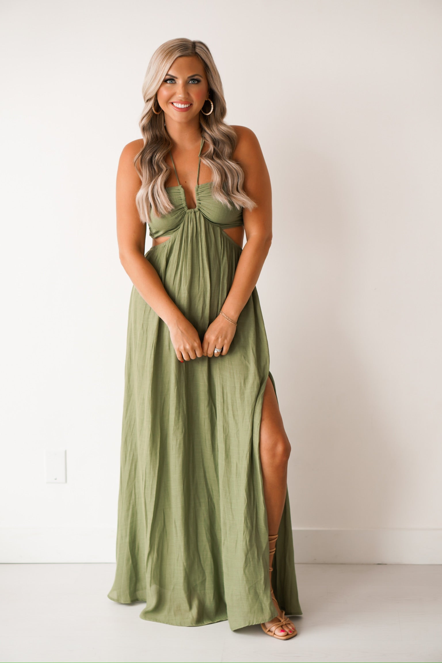 girl standing against a white wall wearing a long green dress that has cutouts on the side of the hips. SHe is wearing sandals
