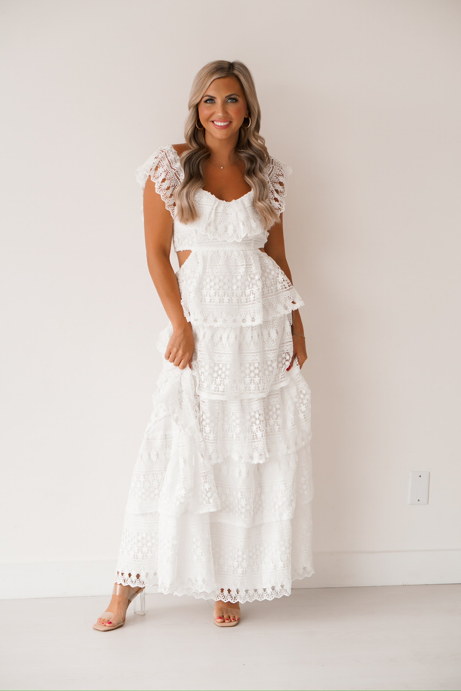 blonde headed lady standing against a white wall wearing an off the shoulder white laced dress