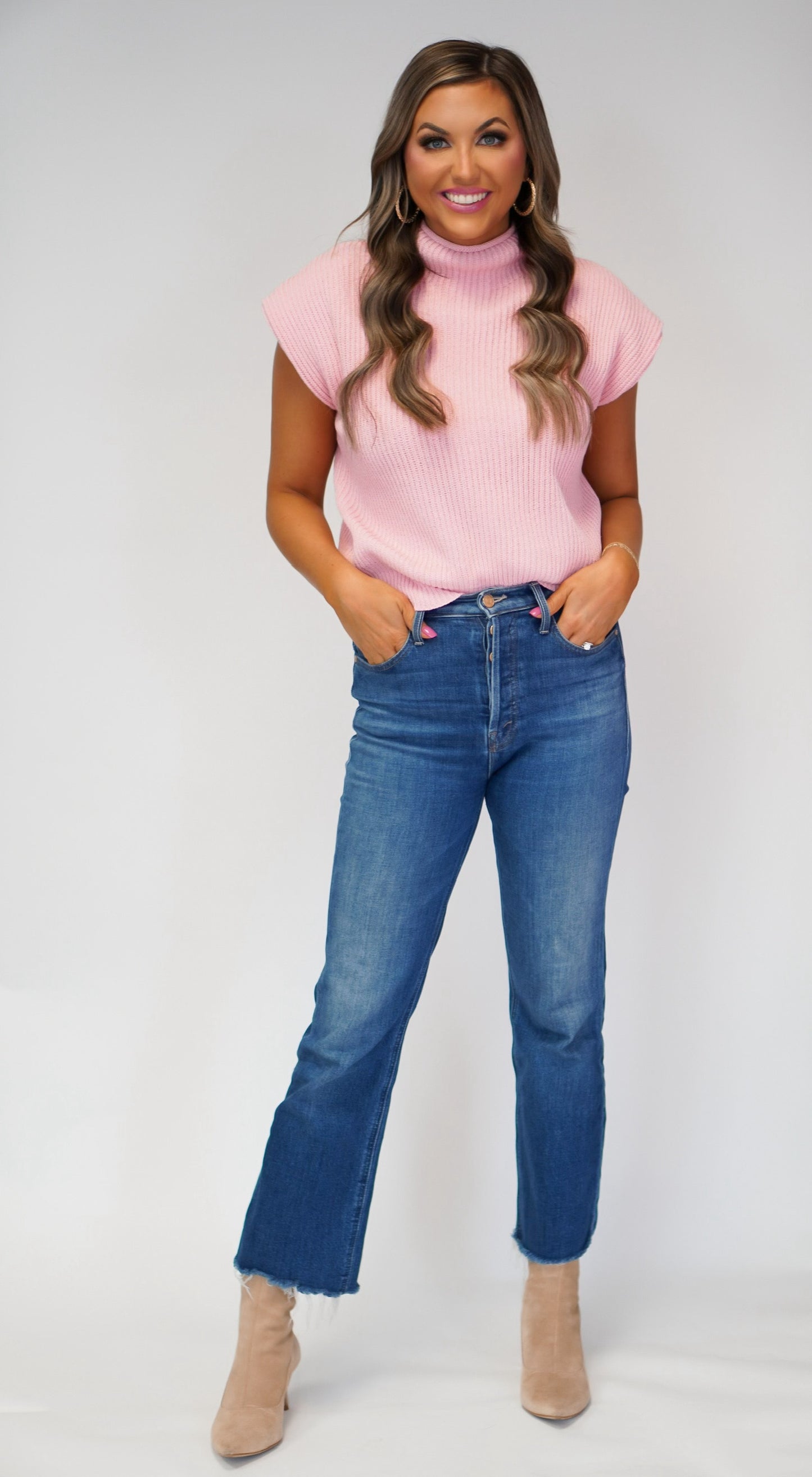 girl standing with hands in jean pockets wearing light pink sweater vest with jeans and boots on 