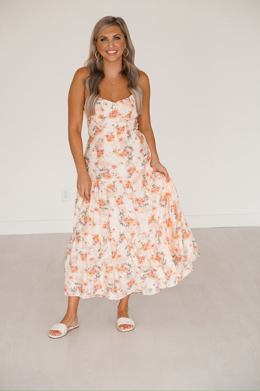 Blonde headed lady standing against a white wall wearing a floral maxi dress with white sandals