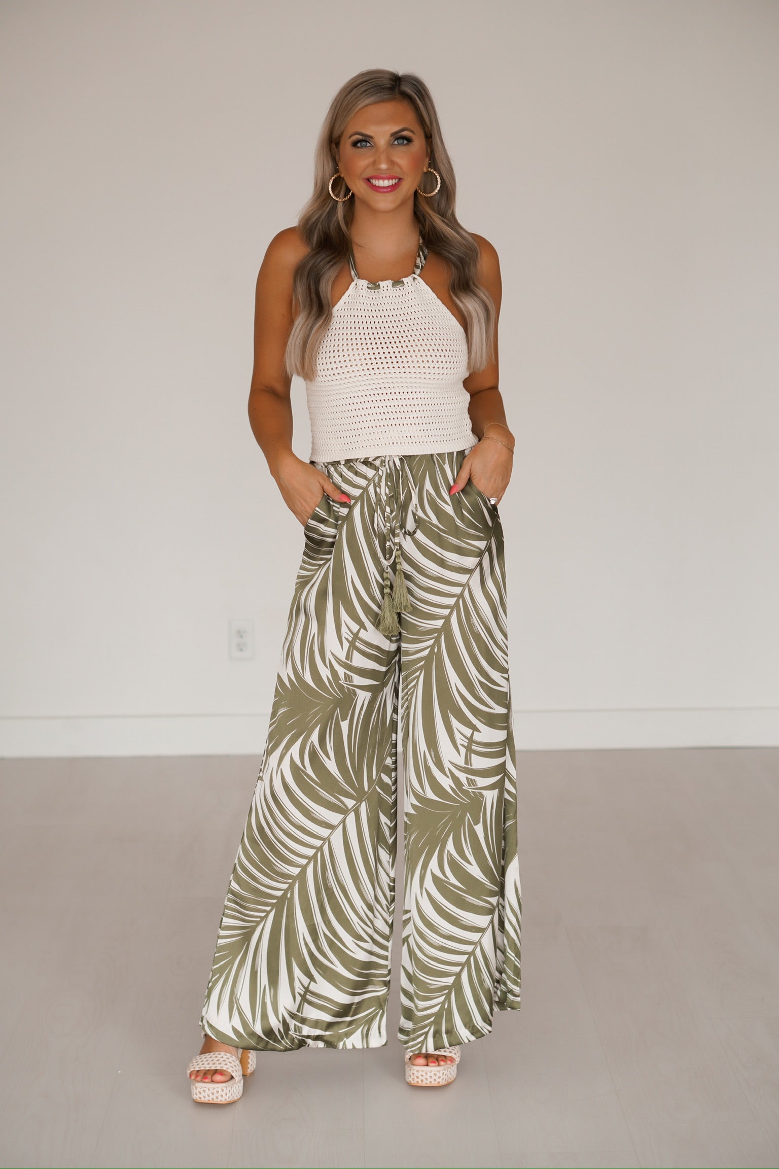 blonde headed girl standing against a white wall wearing pants that have a palm print as well as a knitted top