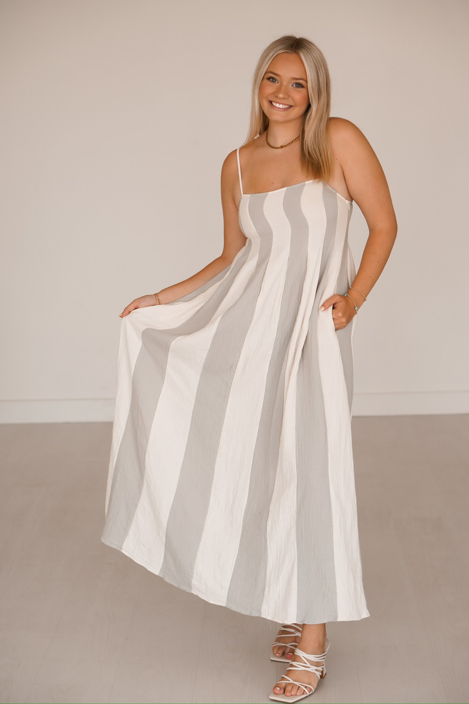 Blonde headed lady standing against a white wall wearing a blue and cream striped dress that has pockets 