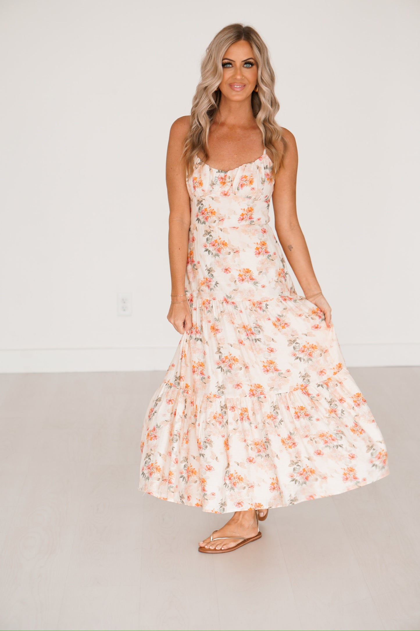 Blonde headed lady standing against a white wall wearing a floral maxi dress with white sandals