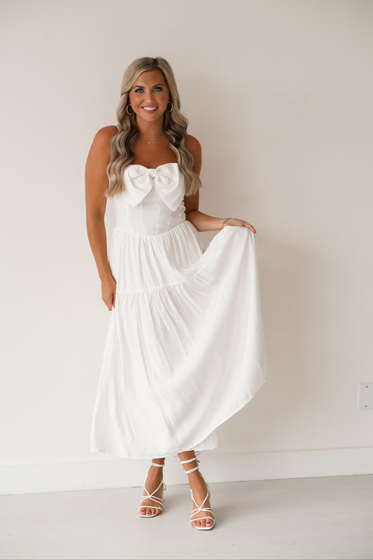 blonde headed lady standing against a white wall wearing a white long dress with a bow on the front. 