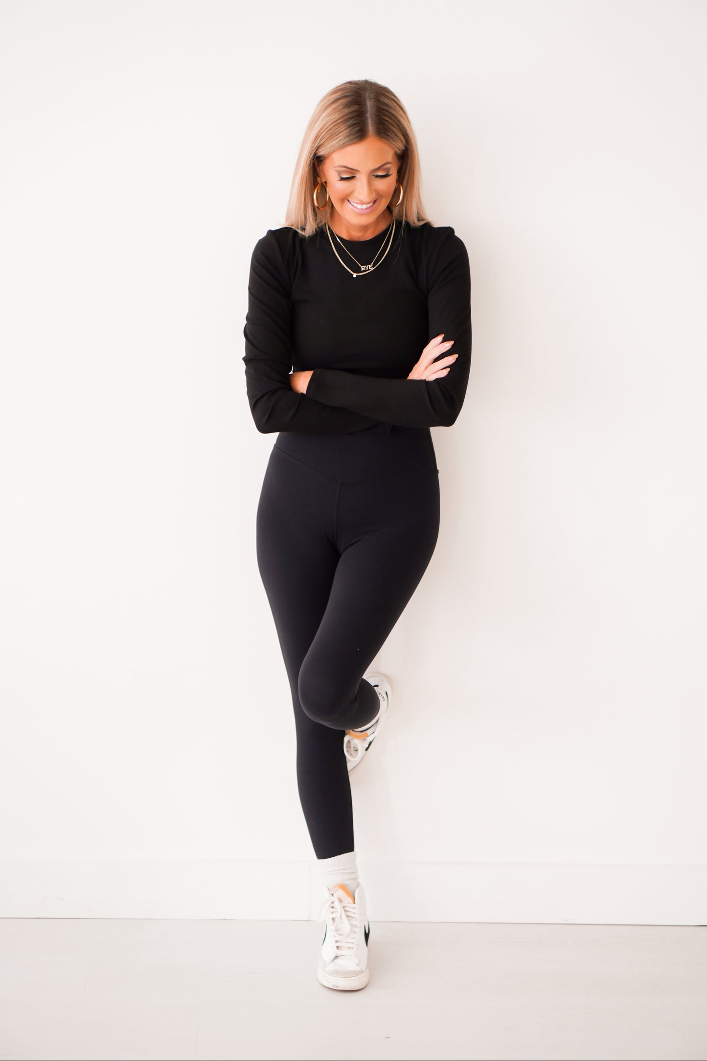 Blonde girl standing in front of white wall wearing black cropped long sleeve ribbed shirt. Her leg is up against the wall. 