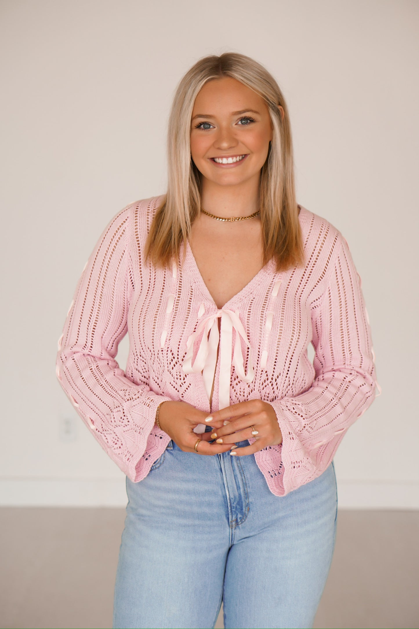 Blonde headed lady standing against a white wall wearing denim jeans and a light pink crocheted top