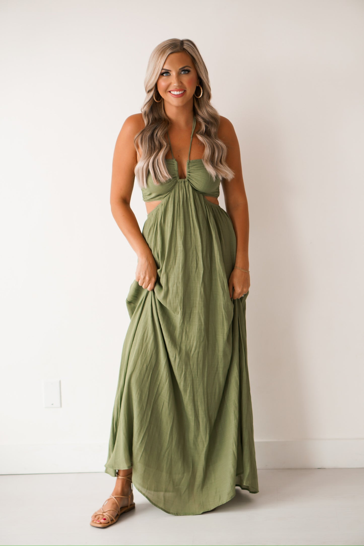 girl standing against a white wall wearing a long green dress that has cutouts on the side of the hips. SHe is wearing sandals