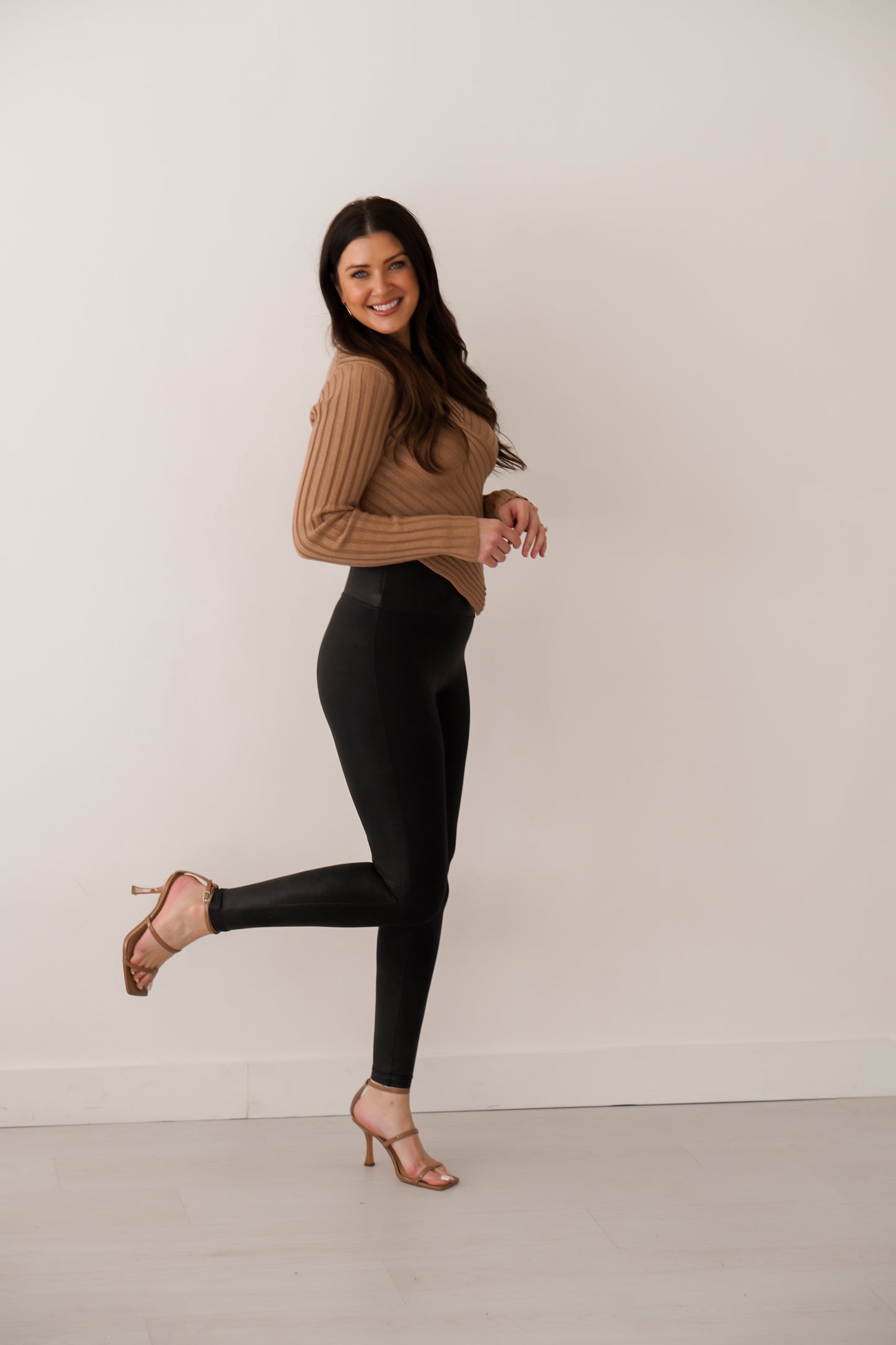 Brunette girl wearing tan sweater with black leggings. She is standing in front of white wall with one leg up with a side view. 