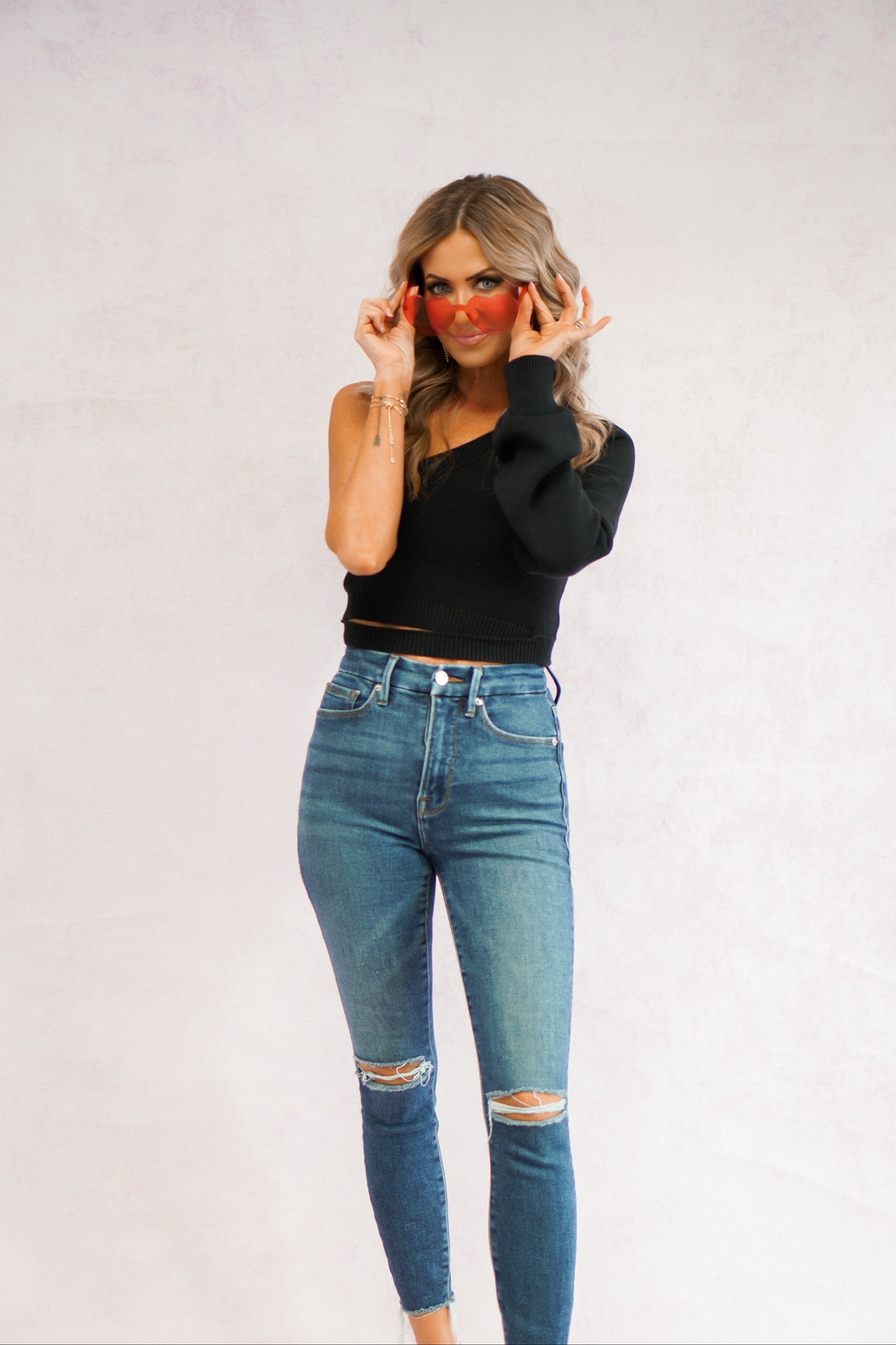 girl showing off her heart shaped glasses while wearing a black one shoulder sweater with cut out detail around the waist and jeans