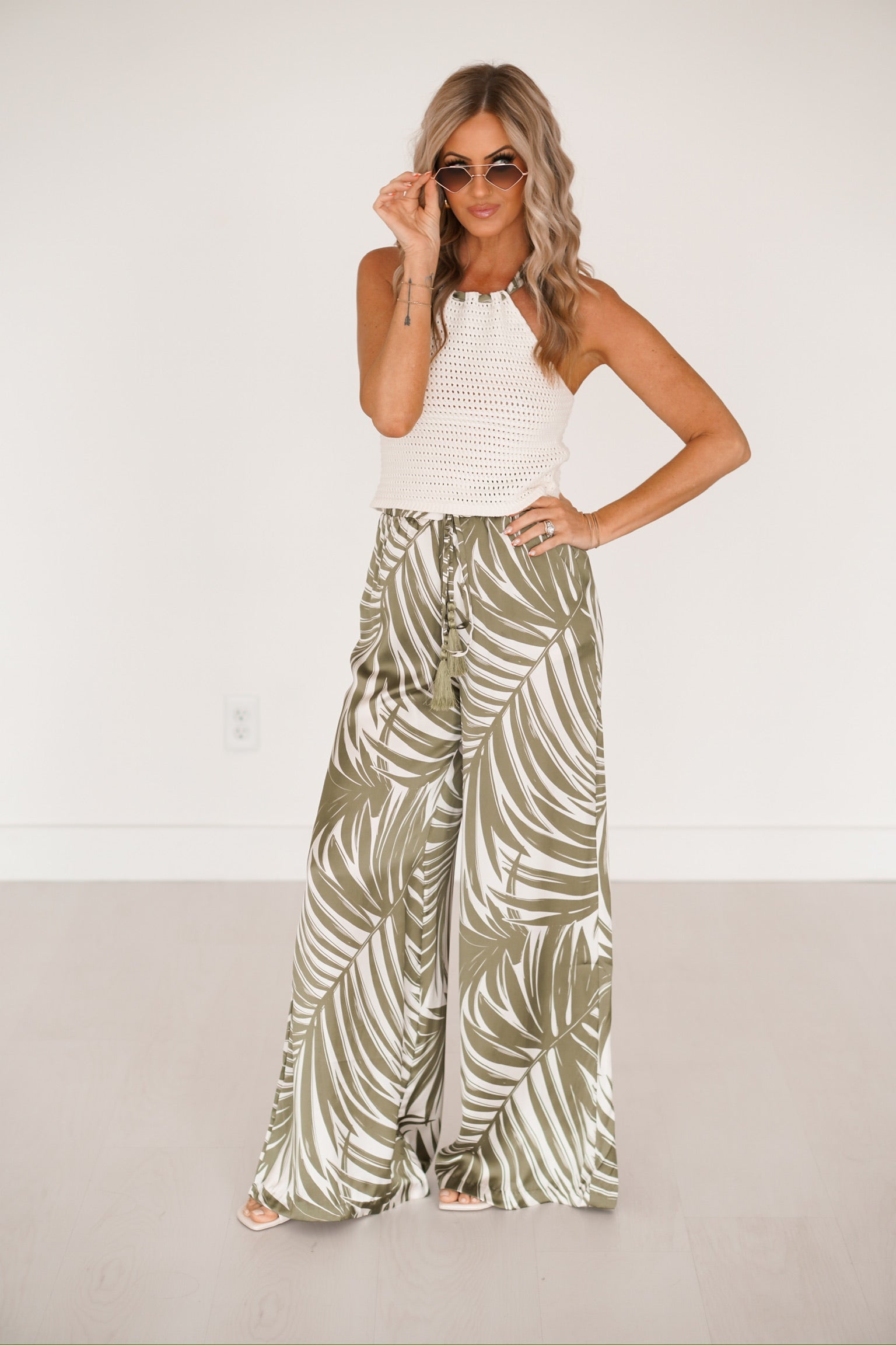 blonde headed girl standing against a white wall wearing pants that have a palm print as well as a knitted top