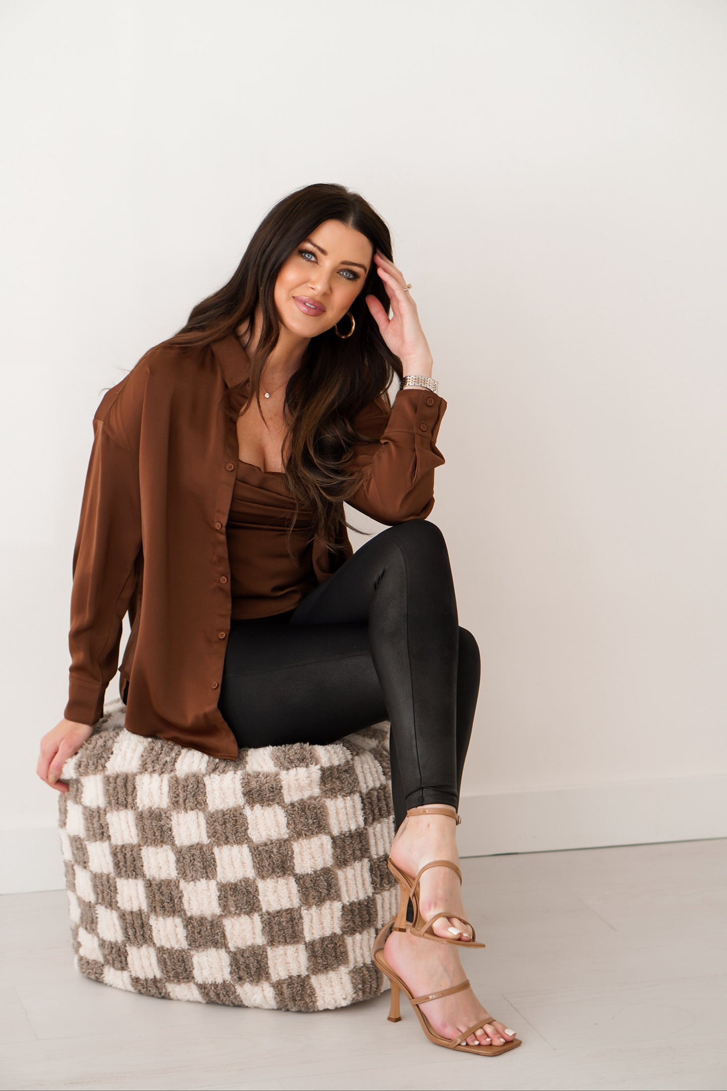 Brunette girl sitting on checkered ottoman wearing chocolate brown silk top set in front of white wall. Her legs are crossed and hand on head. 