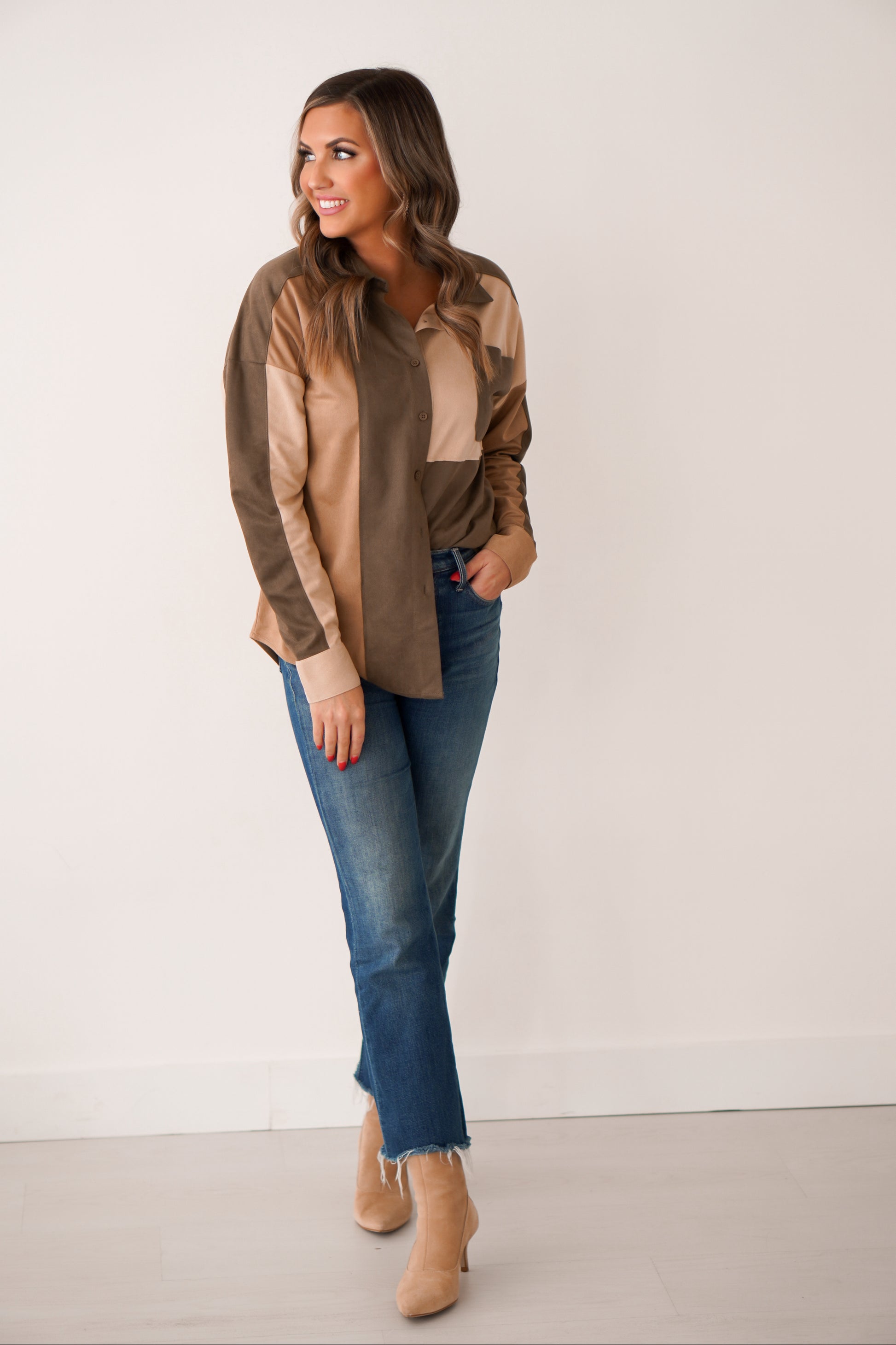 Brunette wearing a color block design shacket in rich tones of olive, brown, and cream. She has it paired with jeans in front of a white wall. 
