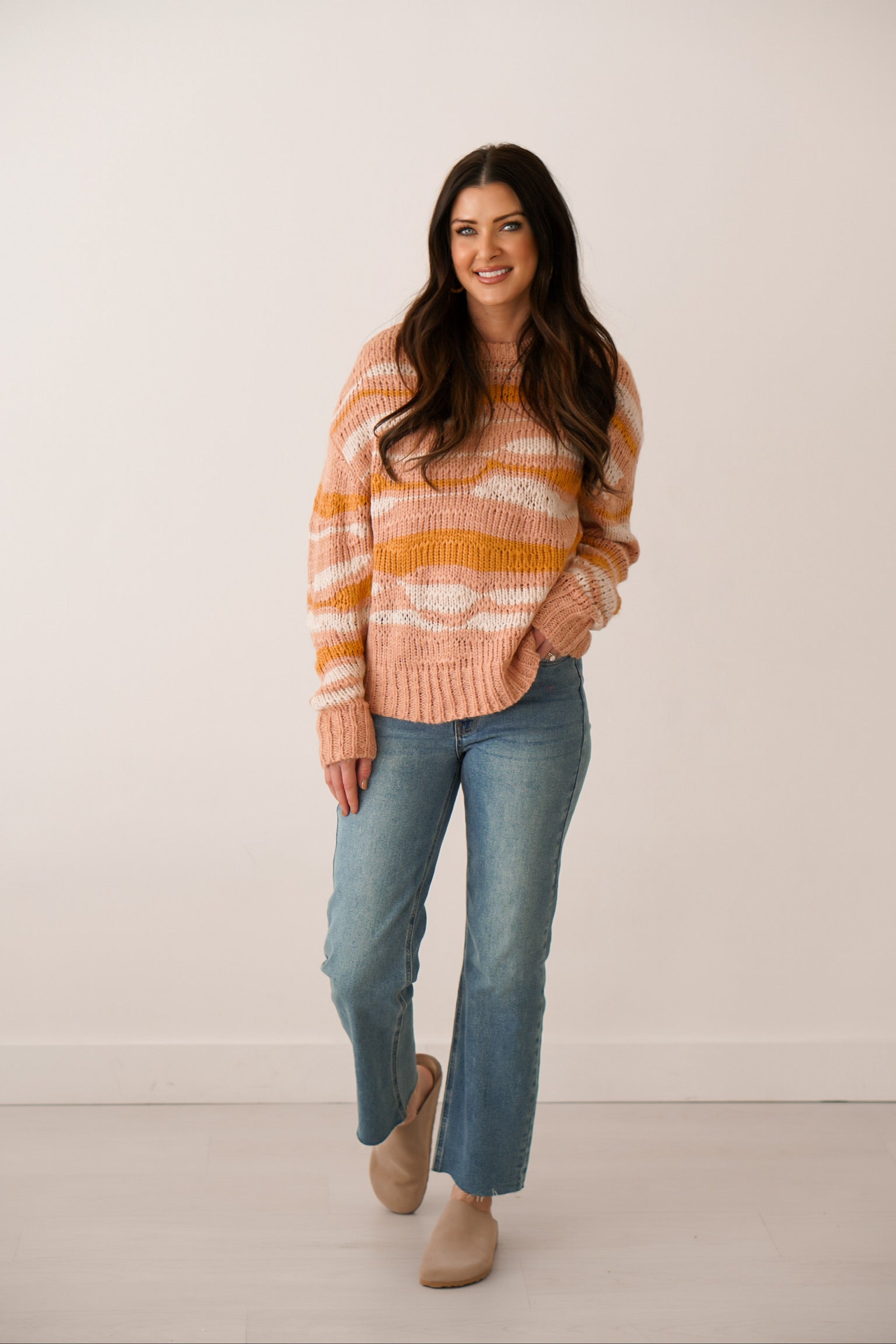 girl standing wearing sweater that is pink white and orange with a wavy landscape design to it with jeans and clogs