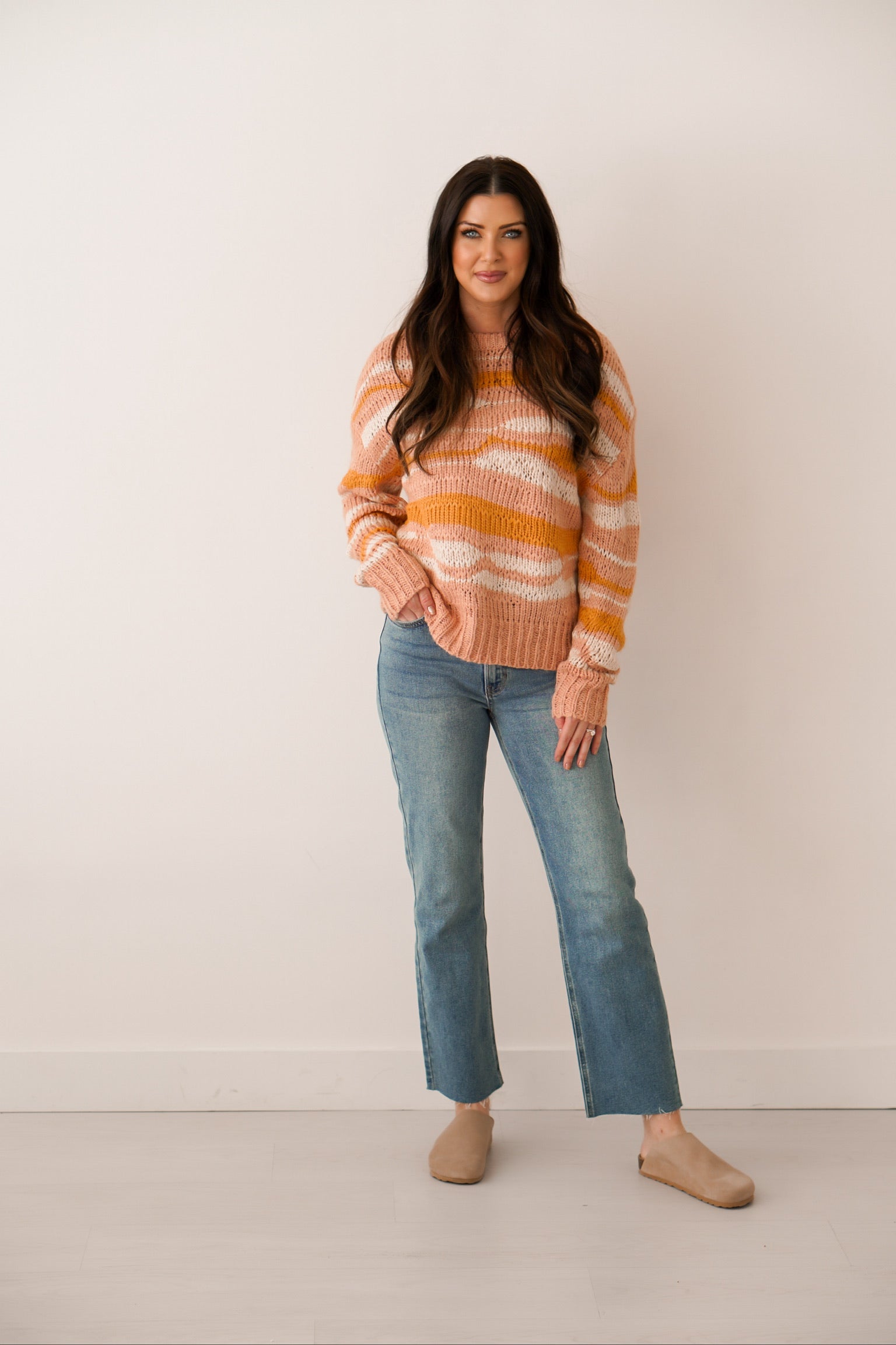 girl standing wearing sweater that is pink white and orange with a wavy landscape design to it with jeans and clogs